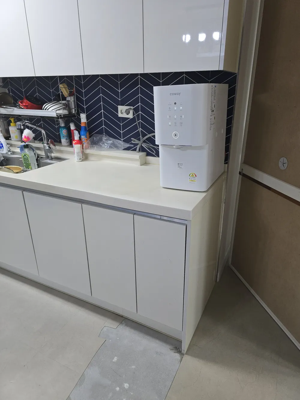 흰색 하부장과 수전, 정수기가 놓인 주방 상판 앞에 바닥이 드러난 모습
